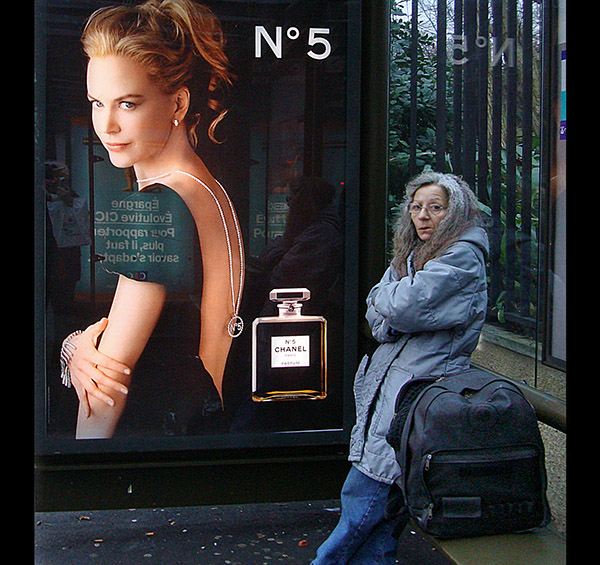 Photo of a grey-haired woman in a bus shelter standing next to a poster of Nicole Kidman for Chanel