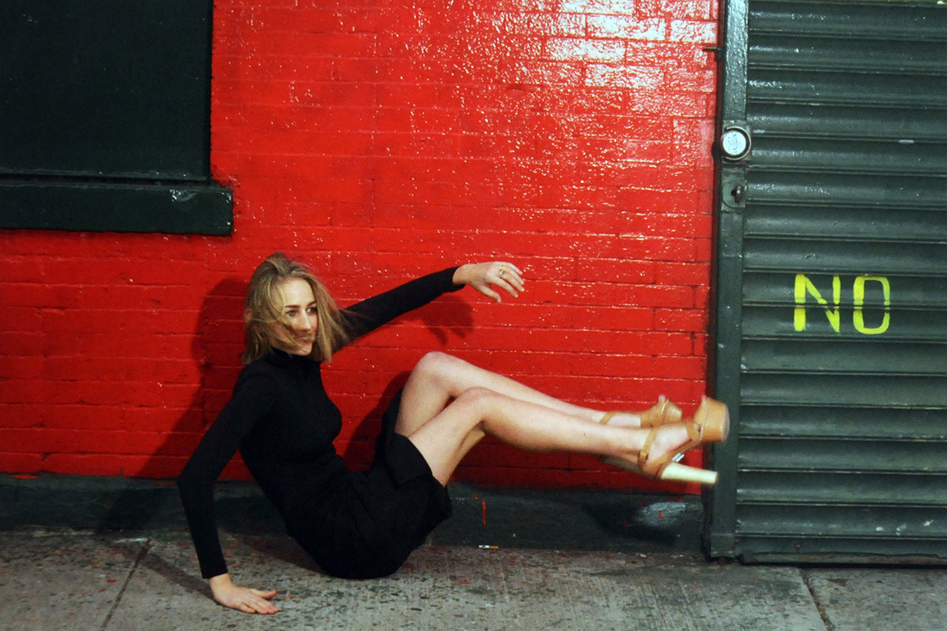 Photo of Vaneeesa Blaylock on her butt with hands and feet in the air, and wearing high platform heels, as though she's just slipped and fallen. Black mini-dress against brightly painted red brick wall