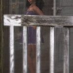 Vaneeesa Blaylock in a mid-thigh purple dress stands behind a screen door in a recreation of an old Tenant Farmer House