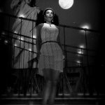 Vaneeesa Blaylock wearing a mid-thigh, sleeveless dress, stands on a rooftop with the Brooklyn Bridge, Statue of Liberty, and a full moon in the sky.