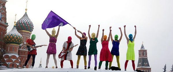 8 members of Pussy Riot stand at a snow covered St. Basil's Cathedral in Russia. Some wave hands in the air. Some play electric guitar.