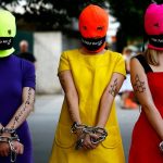 Pussy Riot Solidarity Parade. 3 protestors wear bright dresses and ski masks. Their hands are chained. Their mouths are gagged. They have Amnesty International logos on their arms.