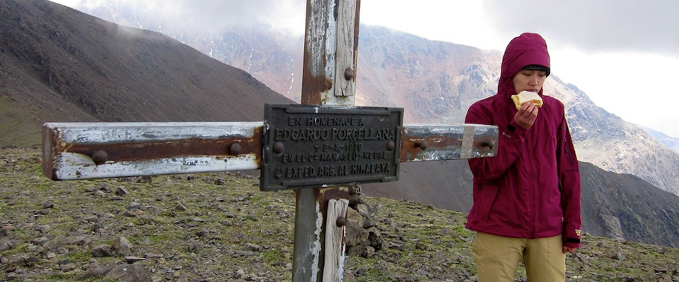 photo of a woman eating a slice of bread on a mountaintop as she walks past a makeshift grave marker in the form of a cross